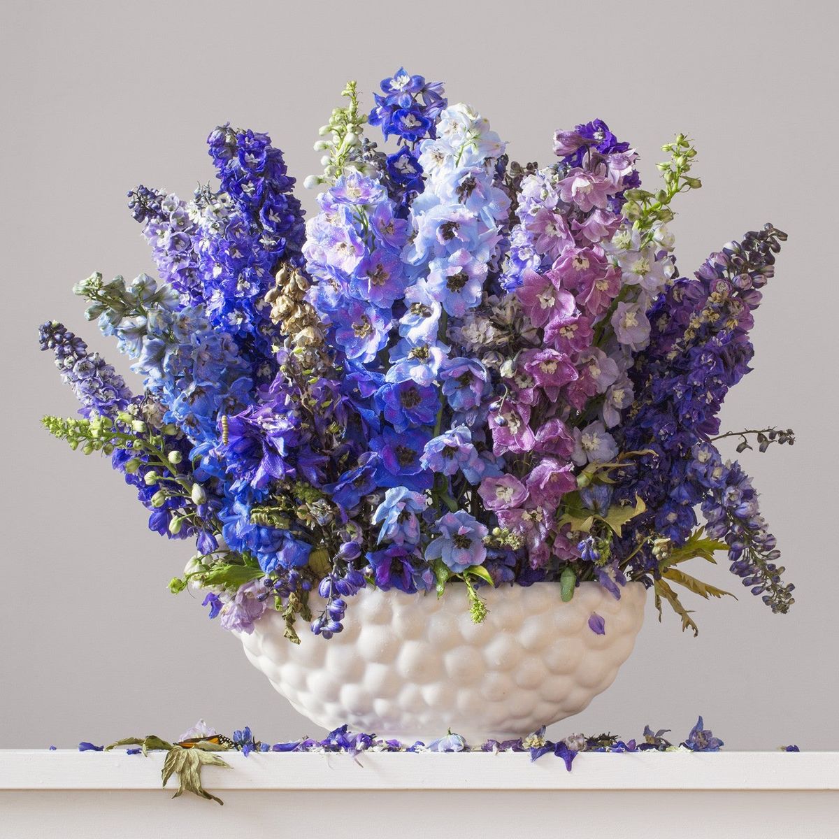 Bouquet of purple, blue, and white flowers in a textured white vase on a light gray background