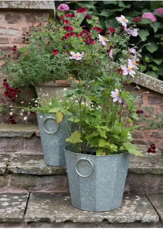 Two galvanized planters with flowers on a stone surface against a brick wall.